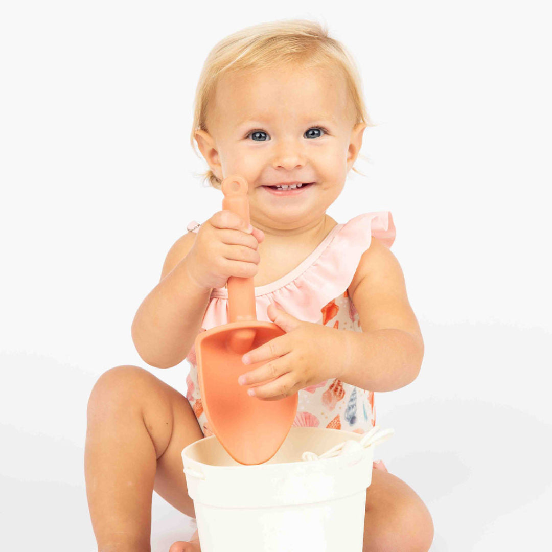 Baby girl smiling sitting down on a white background wearing our sandy Seashell One piece swim suit with ruffles on the light pink truffles. 