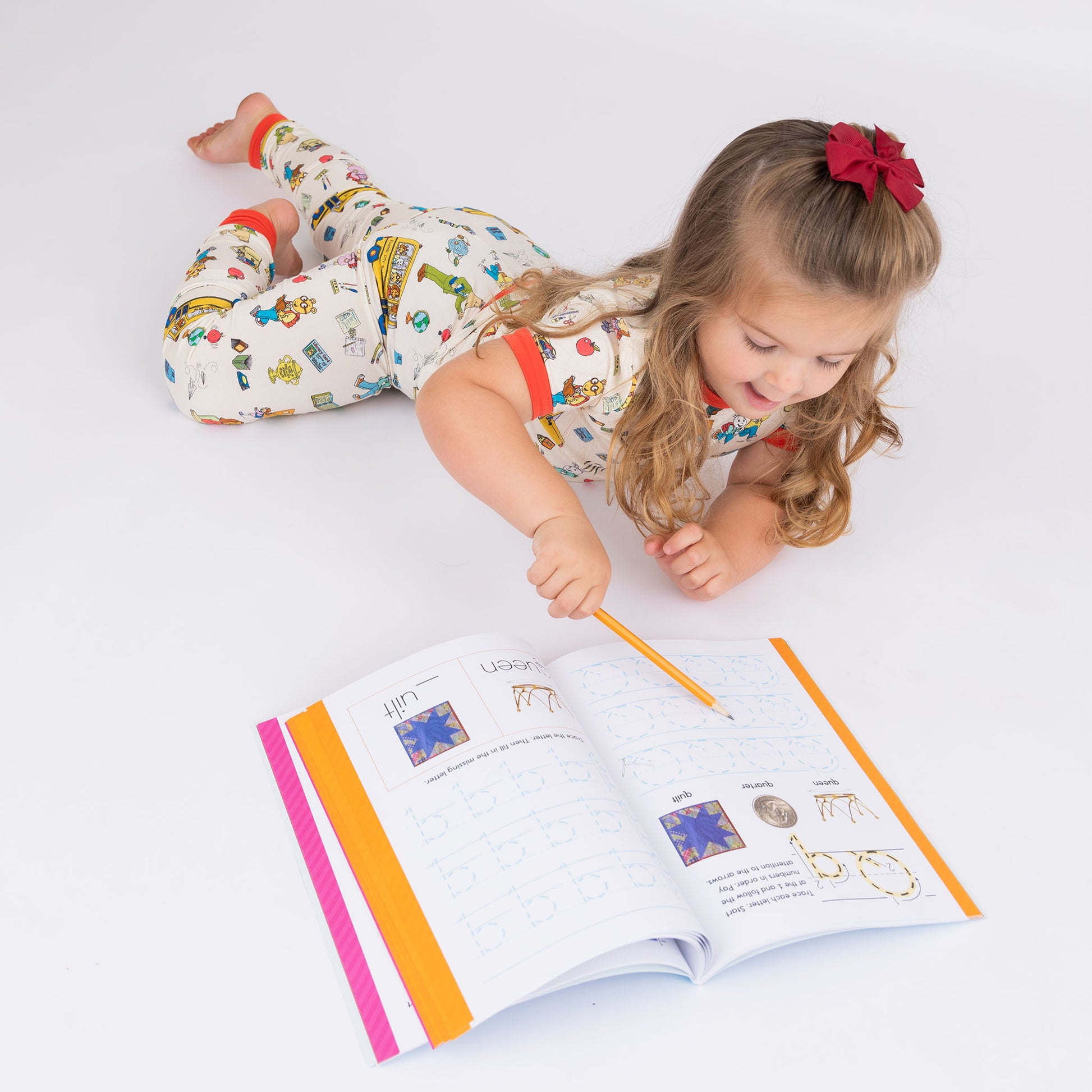 Toddler girl writing on a book wearing our short Sleeve Kids Pajama Pants Set from buttery-soft bamboo, featuring red trims and playful illustrations of Arthur™ PBS Character, books, and school bus, a beige background.
