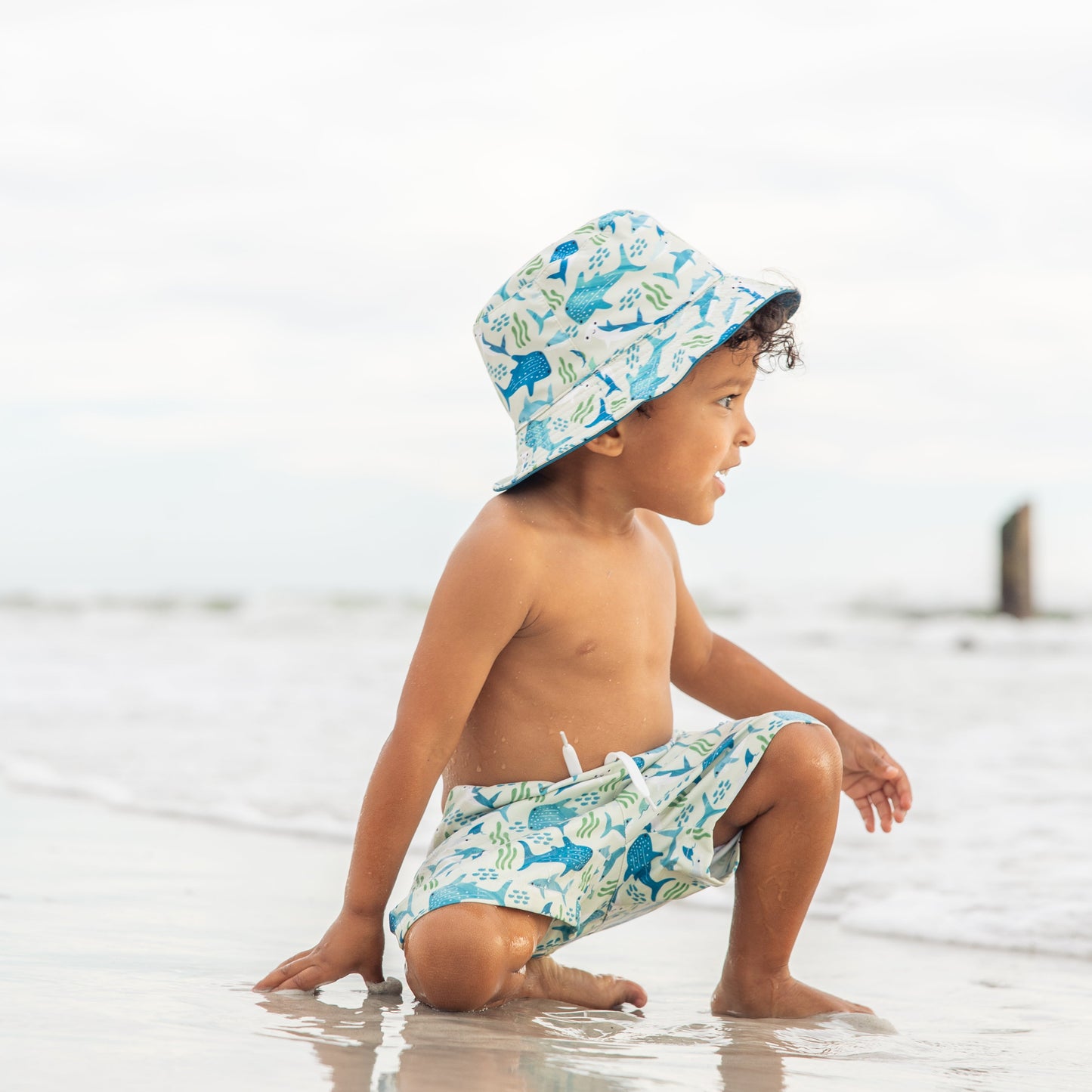 Little boy sitting in the sand wearing the Shark Friends Swim Trunk and Bucket Hat.