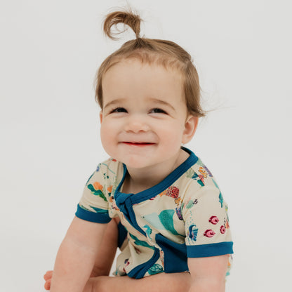 Baby girl wearing our Emerson and Friends Coral Reef Bamboo Baby Shortie Romper, a soft and breathable summer outfit featuring an ocean-inspired print with sharks, sea turtles, and coral, designed for all-day comfort with an easy zip closure.