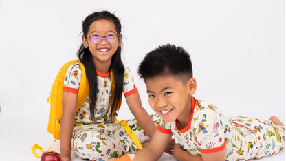 Sibling wearing our short Sleeve Kids Pajama Pants Set from buttery-soft bamboo, featuring red trims and playful illustrations of Arthur™ PBS Character, books, and school bus, a beige background.
