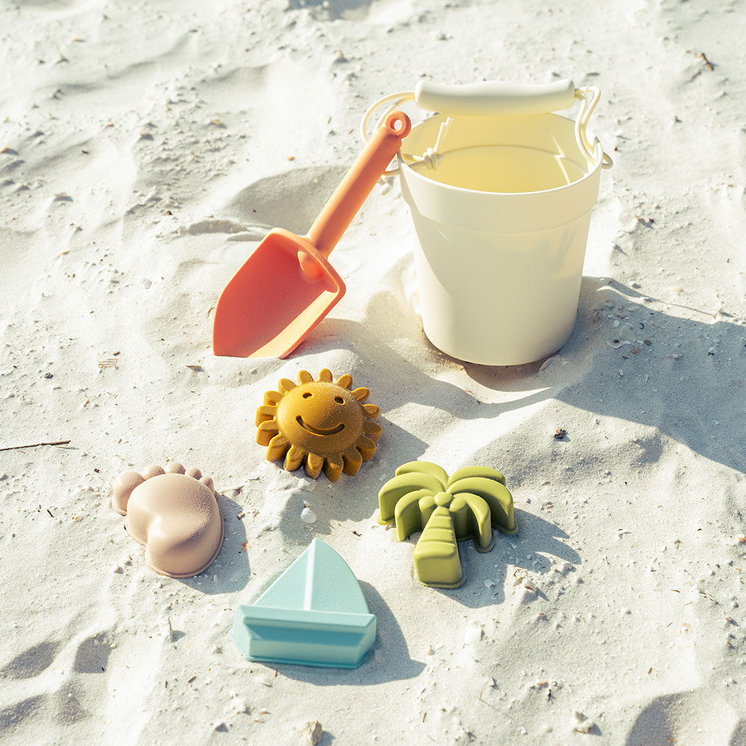 the the silicone white bucket beach toy set. this comes with a pink shovel, green palm tree, blue sailboat, purple footprint, and a yellow sun. this photo was take on the beach the set is spread out on the sand.