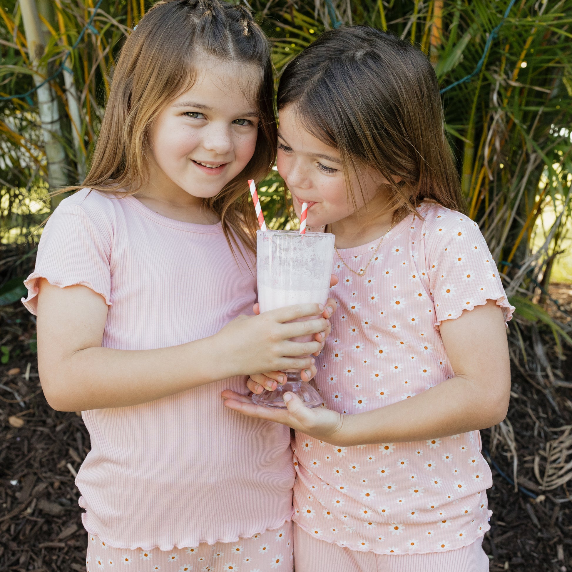 Two young girls in pink outfits drinking from a glass with straws outdoors.
