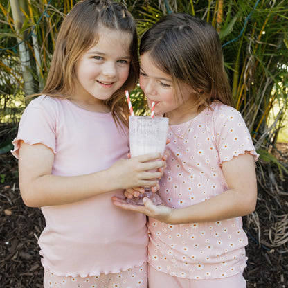 Two young girls in pink outfits drinking from a glass with straws outdoors.