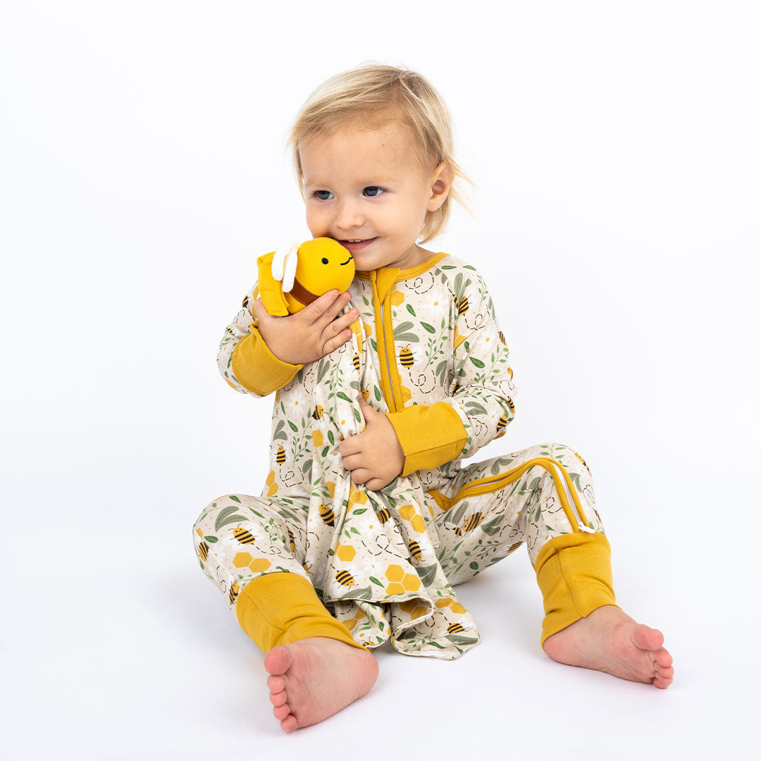 Baby sitting on the floor, wearing our Happy Honey Bee bamboo pajama with mustard yellow trim. The pajama features a bumblebee, honeycomb, and leaf print, paired with a soft lovey/blankie, on a white background.