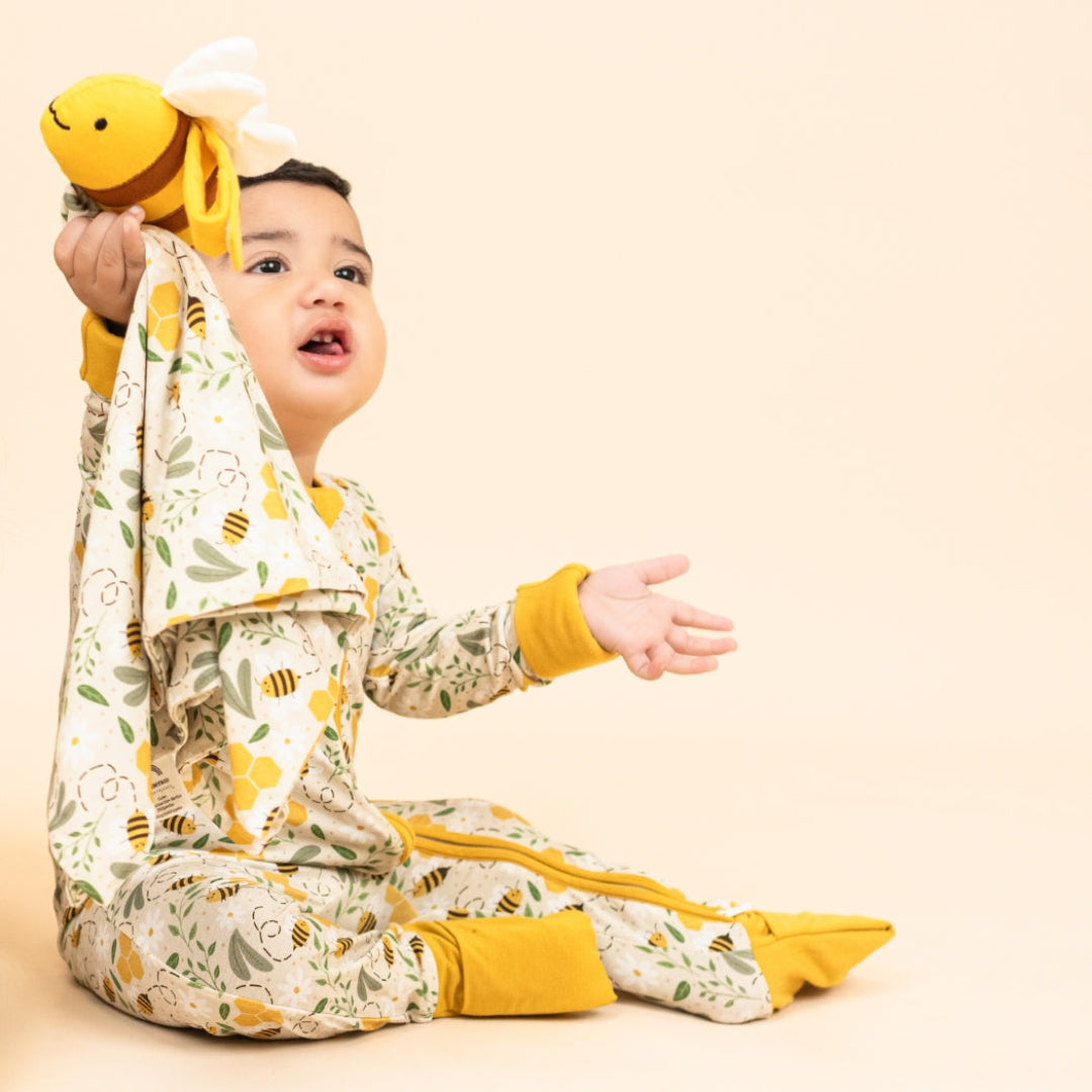 Baby sitting on the floor, wearing our Happy Honey Bee bamboo pajama with mustard yellow trim. The pajama features a bumblebee, honeycomb, and leaf print, paired with a soft lovey/blankie, on a beige background.