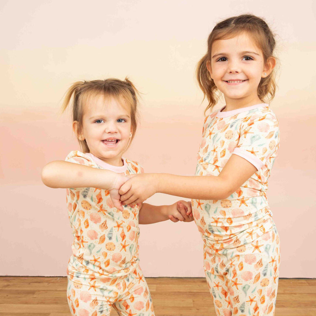 Two girls posing in a pink and yellow gradient background wearing our 2 piece bamboo pajama from our Sandy Seashell collection. 