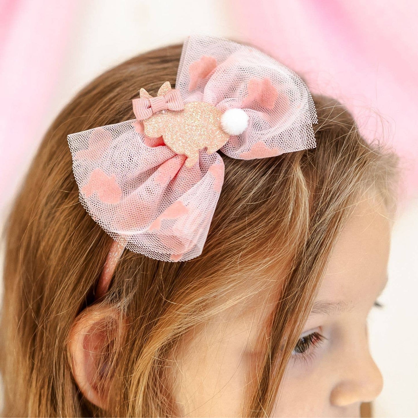 Child wearing a pink hair bow with a gold center against a blurred pink background