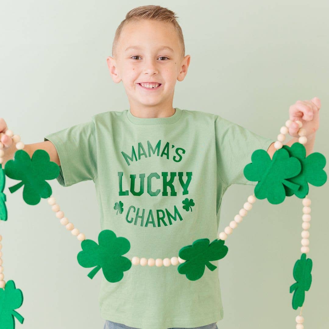 Child wearing a green shirt with 'Mama's Lucky Charm' text, holding a shamrock necklace.