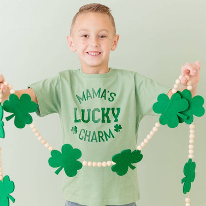 Child wearing a green shirt with 'Mama's Lucky Charm' text, holding a shamrock necklace.