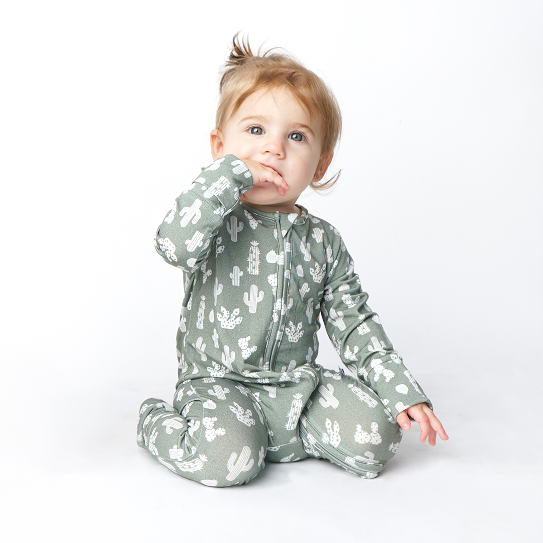 young toddler girl sits on her knees with her hand in her mouth. she is wearing the "stay sharp" baby convertible. the "stay sharp" print is a variety of different white cacti on a greyish/green background. 