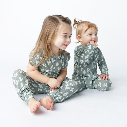 2 girls sit next to one another on the floor. the girl on the left wears the "stay sharp" matching pajama set. the girl on the left she is wearing the "stay sharp" baby convertible. the "stay sharp" print is a variety of different white cacti on a greyish/green background. 
