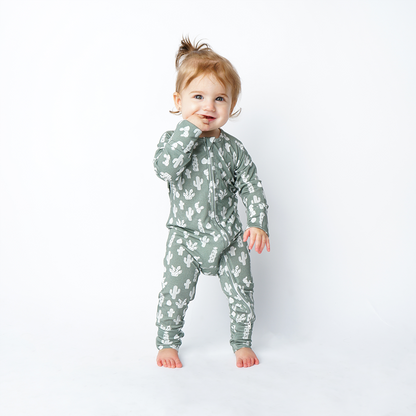 young toddler girl smiles with her hand in her mouth. she is wearing the "stay sharp" baby convertible. the "stay sharp" print is a variety of different white cacti on a greyish/green background. 