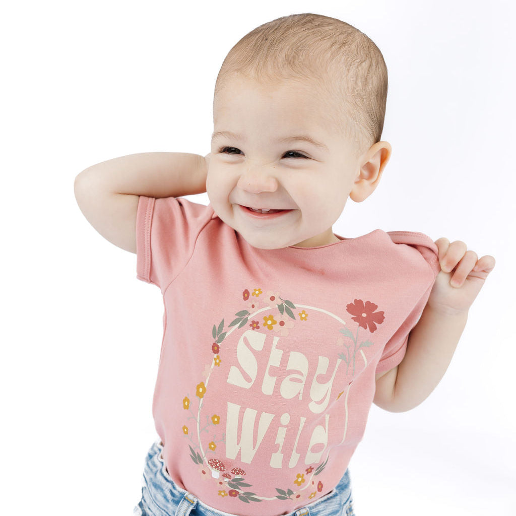 A baby wearing a pink short-sleeve t-shirt with a floral graphic and the words 'Stay Wild' in white, paired with light blue denim jeans, posing against a solid white background.