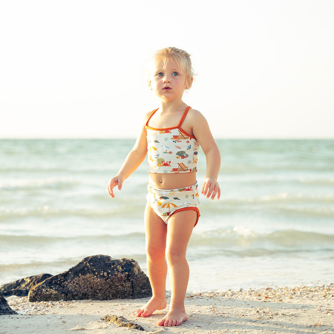 young girl stands in the sand in her "beach days" reversible bikini. the "beach days" print is a scattered print of beach umbrellas, beach balls, sand buckets and shovels, coral, shells, beach chairs, seagulls, flipflops, and sunglass mixed in a orderly and creative way. there is a rock and the ocean in the background. 