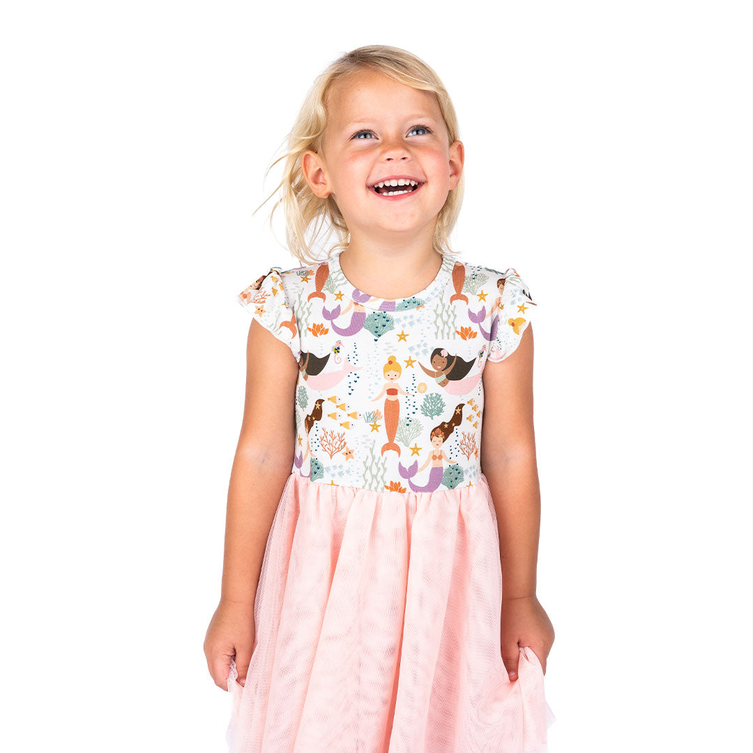 a young girl smiles happily in her tulle twirl dress in the making waves print. the "making waves" print has a diverse spread of mermaids, sea coral, starfish, fish, and bubbles all spread out in different colors. this is all put on a beige background.