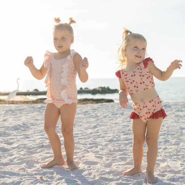 Two young girls in swimsuits standing on a sandy beach with ocean in the background.