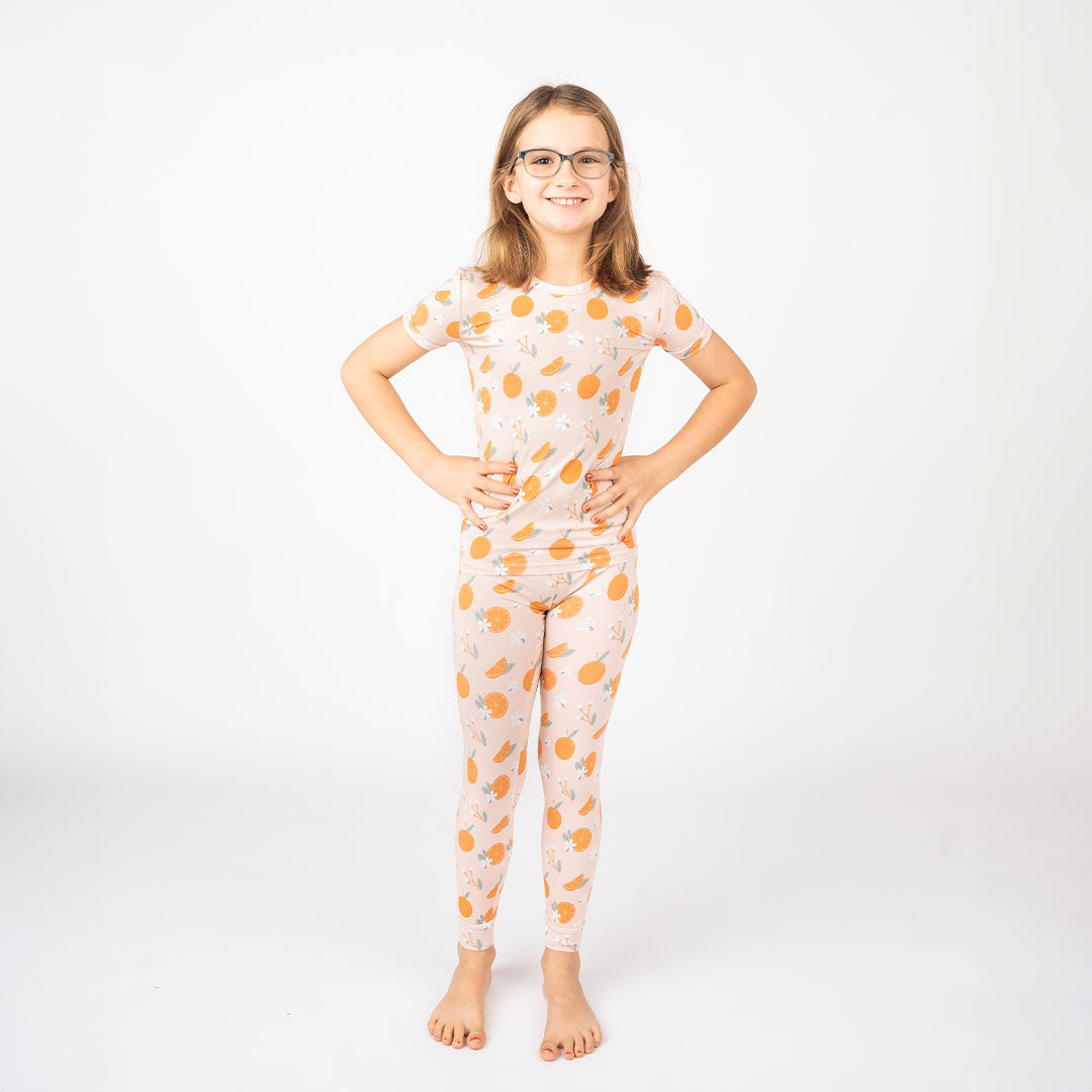a young girl poses with her hands on her hips. she is wearing the 2-piece matching pajama set in the "freshly squeezed" print. the "freshly squeezed" print has an assortment of full and half cut oranges scattered around. there is also flower heads and flower stems that intermingle within the print. this is all space out around a pink background space. 