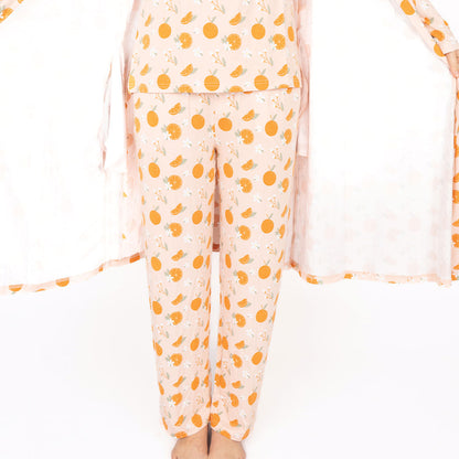 someone posing in the "freshly squeezed" bamboo lounge relaxed pants. they are also wearing the freshly squeezed women's top and spreading open their "freshly squeezed" robe to show her clothes underneath. the "freshly squeezed" print has an assortment of full and half cut oranges scattered around. there is also flower heads and flower stems that intermingle within the print. this is all space out around a pink background space. 