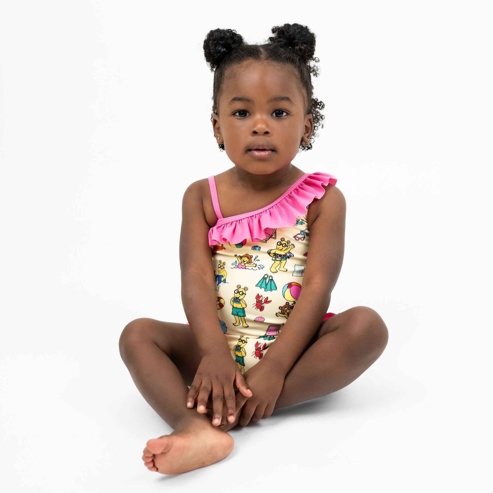 Toddler sitting down with our Emerson and Friends Arthur's Family Vacation Girls One-Piece Swimsuit, featuring a playful beach-themed print with Arthur and friends, ruffle details on the shoulder and legs, and a soft, stretchy fabric for a fun and comfortable swimwear option.