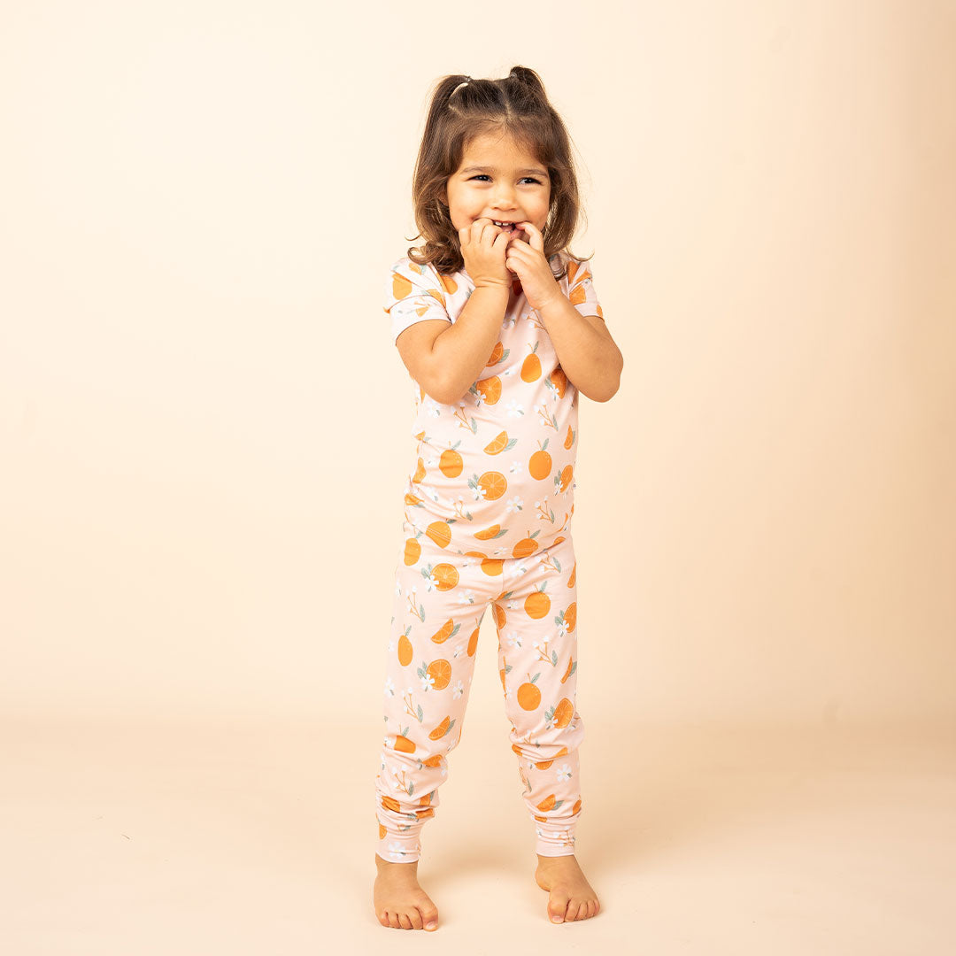 a cute toddler girl stand with her fingers covering her mouth while she smiles big. she is wearing the "freshly squeezed" 2-piece matching pajama set. the "freshly squeezed" print has an assortment of full and half cut oranges scattered around. there is also flower heads and flower stems that intermingle within the print. this is all space out around a pink background space. 
