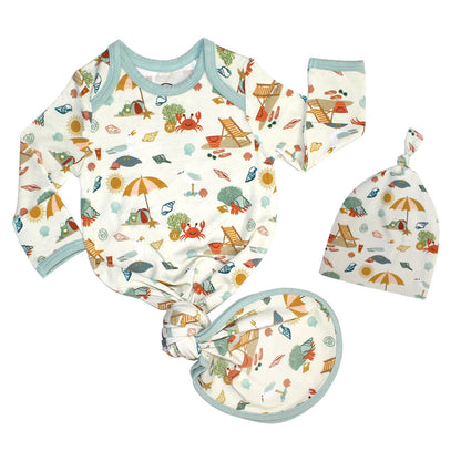 the "beach days" gown and hat set. the gown ties in a knot at the bottom. the "beach days" print is a scattered print of beach umbrellas, beach balls, sand buckets and shovels, coral, shells, beach chairs, seagulls, flipflops, and sunglass mixed in a orderly and creative way. 