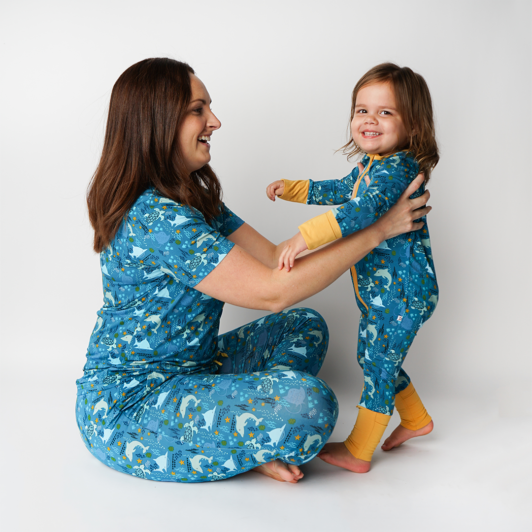 mom sits criss-cross on the floor smiling at her toddler girl. the toddler stands in her "ocean friends" convertible. the "ocean friends" print is a combination of dolphins, stingrays, fish, starfish, coral, bubbles, and sharks, all spread out on a deep sea blue background. 