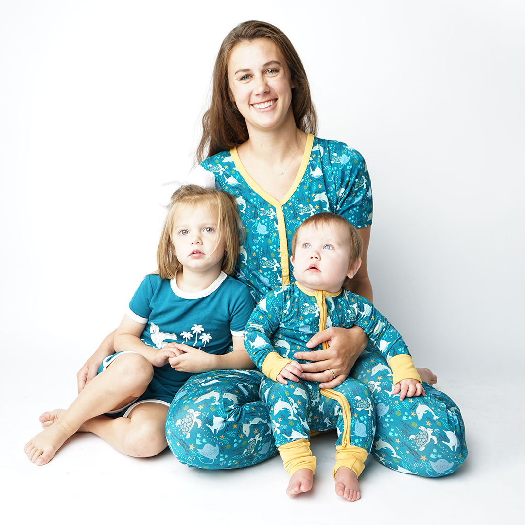 mom sits on the floor smiling in her "ocean friends" women's top and joggers. the baby sits on her right in the "ocean friends convertible. and a toddle sits on her left in the "terry ocean blue" shorts and the "just beachy" tee. the "ocean friends" print is a combination of dolphins, stingrays, fish, starfish, coral, bubbles, and sharks, all spread out on a deep sea blue background. 