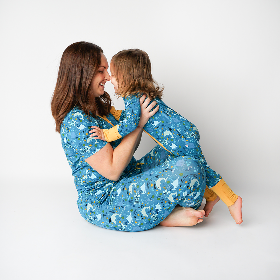 mom sits criss-cross on the floor with her nose presses up against her daughters nose. the picture is taken from the side and we see them both wearing "ocean friends" prints. the mom in the women's top and jogger bottoms, and the baby in the convertible. the "ocean friends" print is a combination of dolphins, stingrays, fish, starfish, coral, bubbles, and sharks, all spread out on a deep sea blue background. 