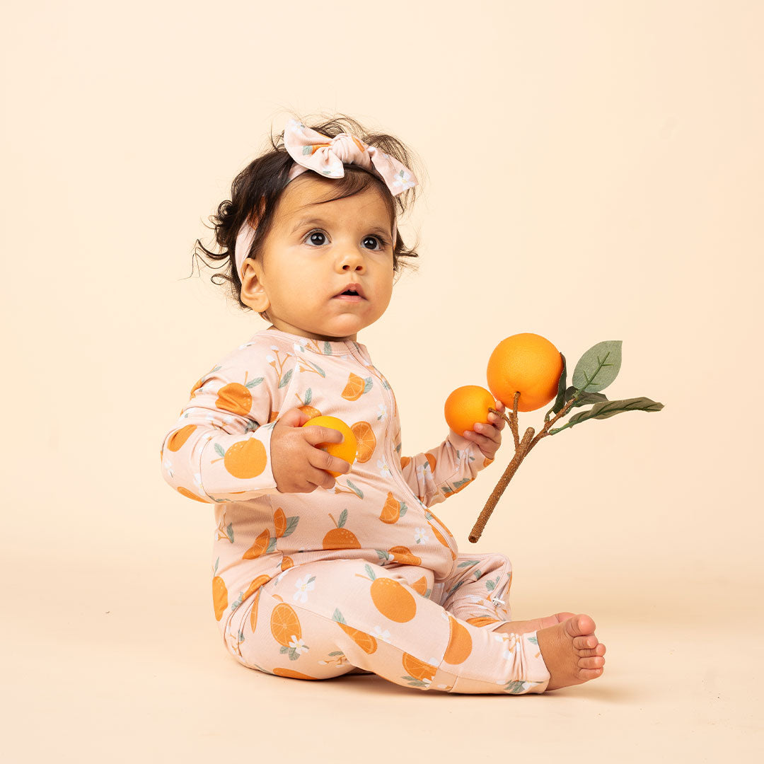 cute baby sitting on the floor holding a couple of oranges. she is wearing the "freshly squeezed" convertible and the headband. the "freshly squeezed" print has an assortment of full and half cut oranges scattered around. there is also flower heads and flower stems that intermingle within the print. this is all space out around a pink background space. 