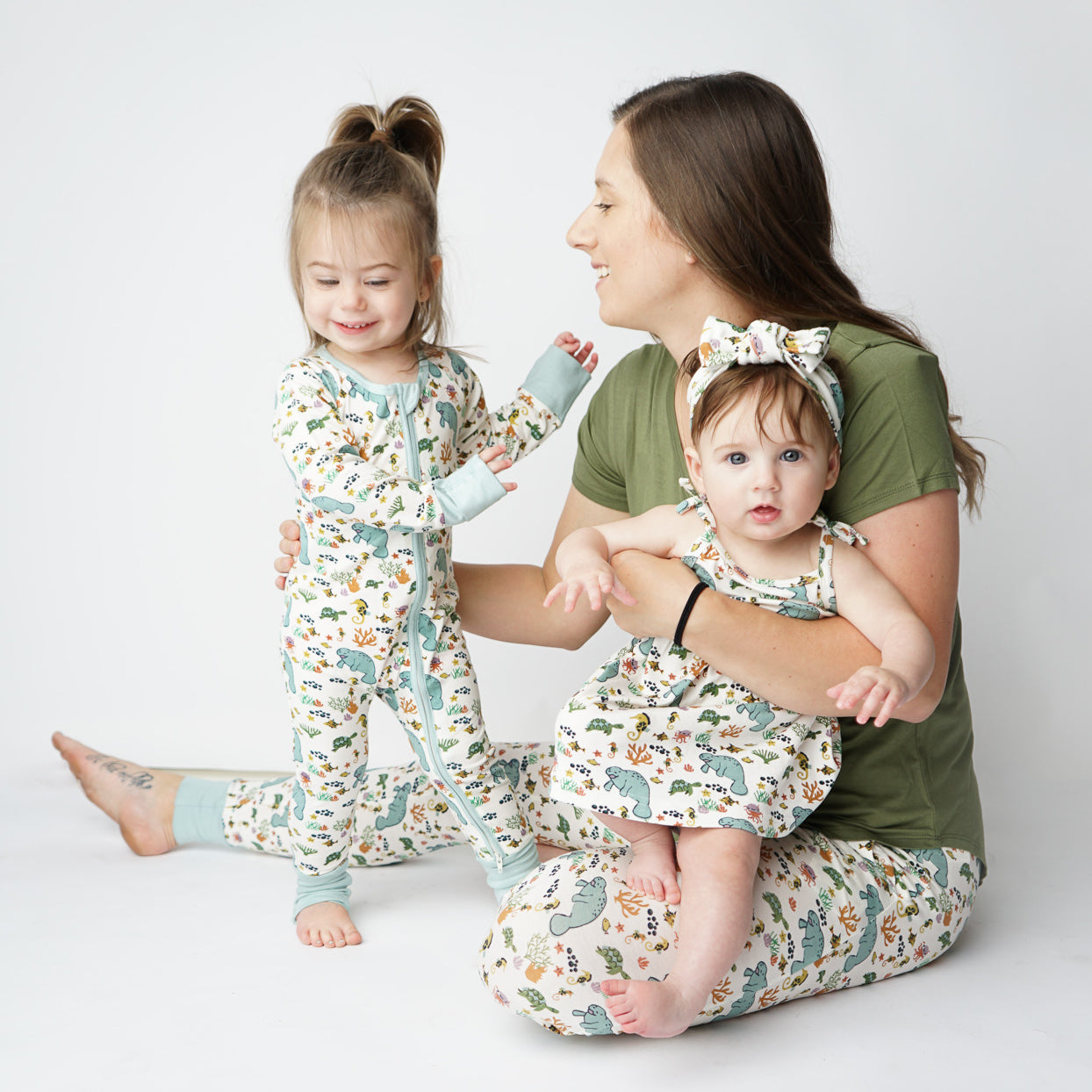 a mom with her 2 daughters playing on the floor. the baby sits in her mom's lap wearing the manatee sundress and headband. the daughter stands next to mom wearing the manatee 2-piece matching pajama set. the mom wears the manatee joggers and the "olive green" women's top.