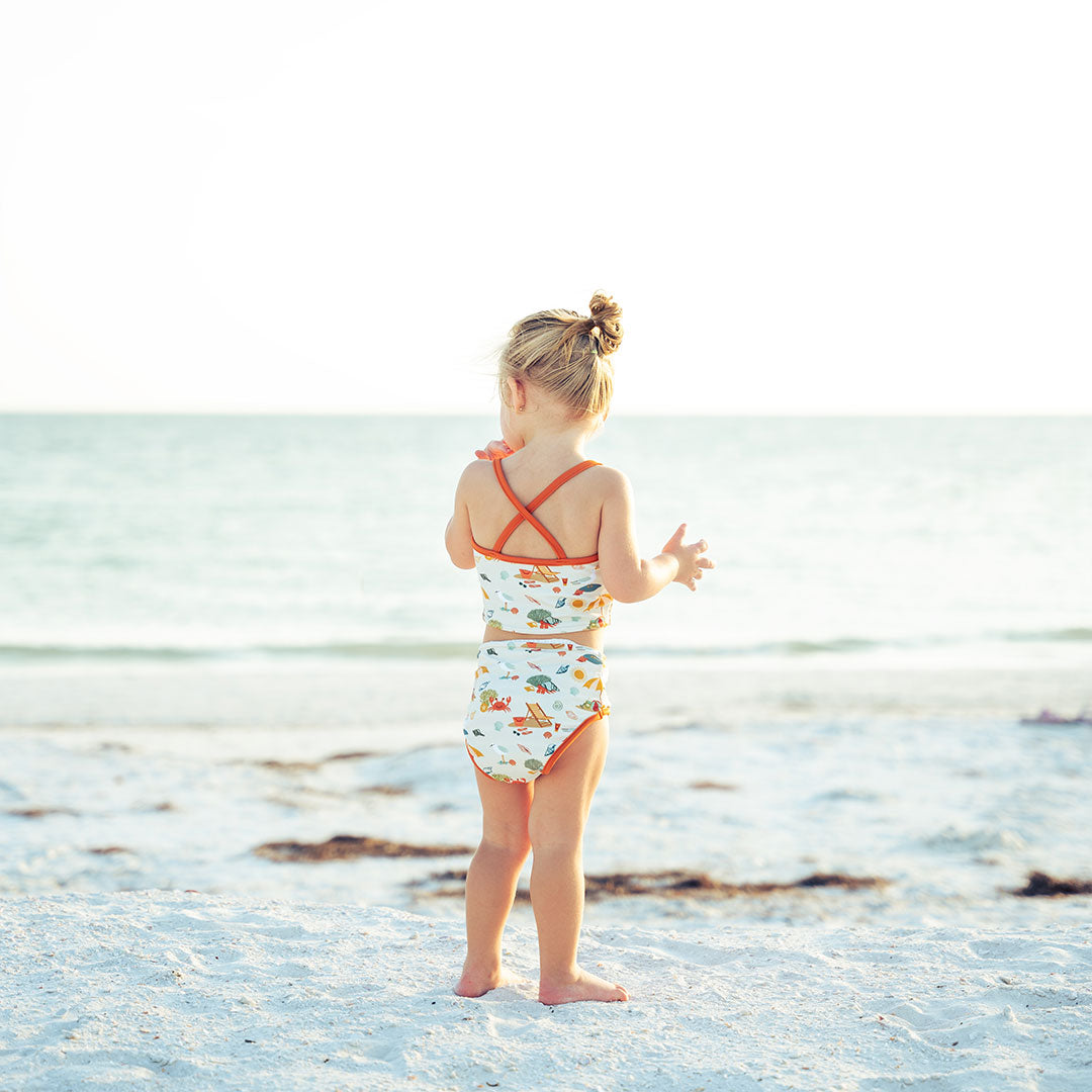young girl stands with her back to the camera on the beach. she is looking at the ocean. she wears the "beach days" reversible bikini. the "beach days" print is a scattered print of beach umbrellas, beach balls, sand buckets and shovels, coral, shells, beach chairs, seagulls, flipflops, and sunglass mixed in a orderly and creative way. 
