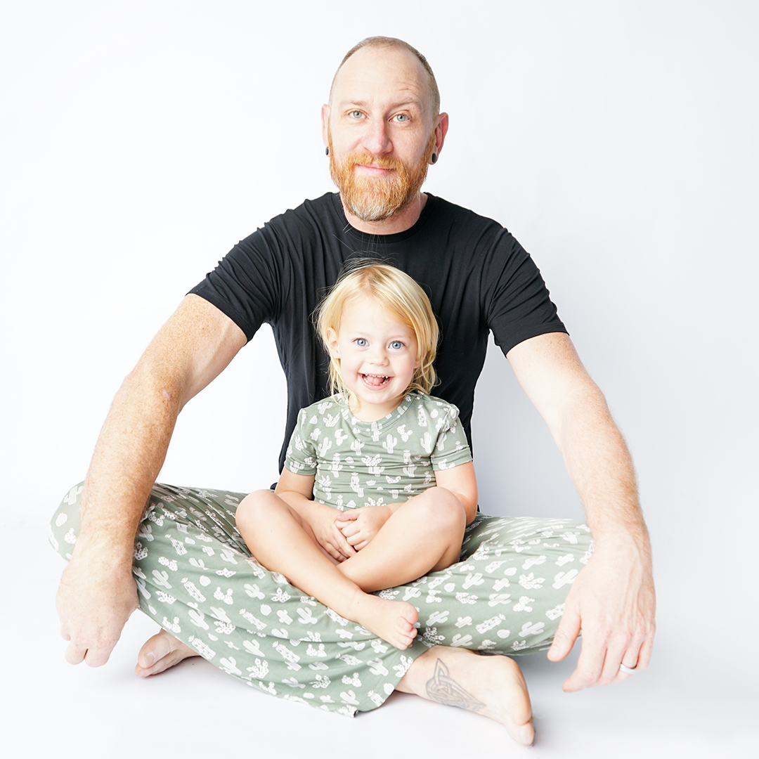 dad sits criss-cross on the floor with his daughter in his lap they are both wearing "stay sharp" prints. dad in the relaxed pants and the girl in the short sleeved matching pajama set. v