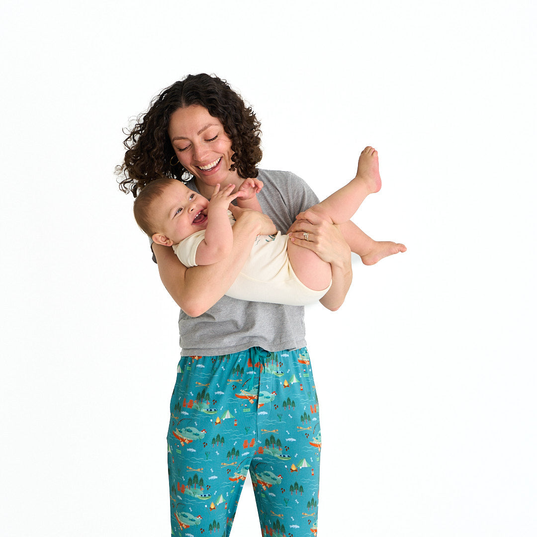 mom wears the "jump in the lake" relaxed pants and hold her baby. the "jump in the lake" pattern is a print to capture the day in the life on a lake. you can fine fishing poles, tents, camp fires, adirondack chairs, marsh mellows, ores, canoes, forest trees, and different types of fish and fishing gear.