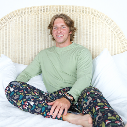 a guy sits on a white bed wearing the "night forest" relaxed pants the "night forest" print is a night time themed design. you can see an array of forest animals ranging from, deer, bears, bunnies, birds, forest trees and leaves, flowers, and mushrooms. there are also starts and moons scattered around to enhance the night time atmosphere. 