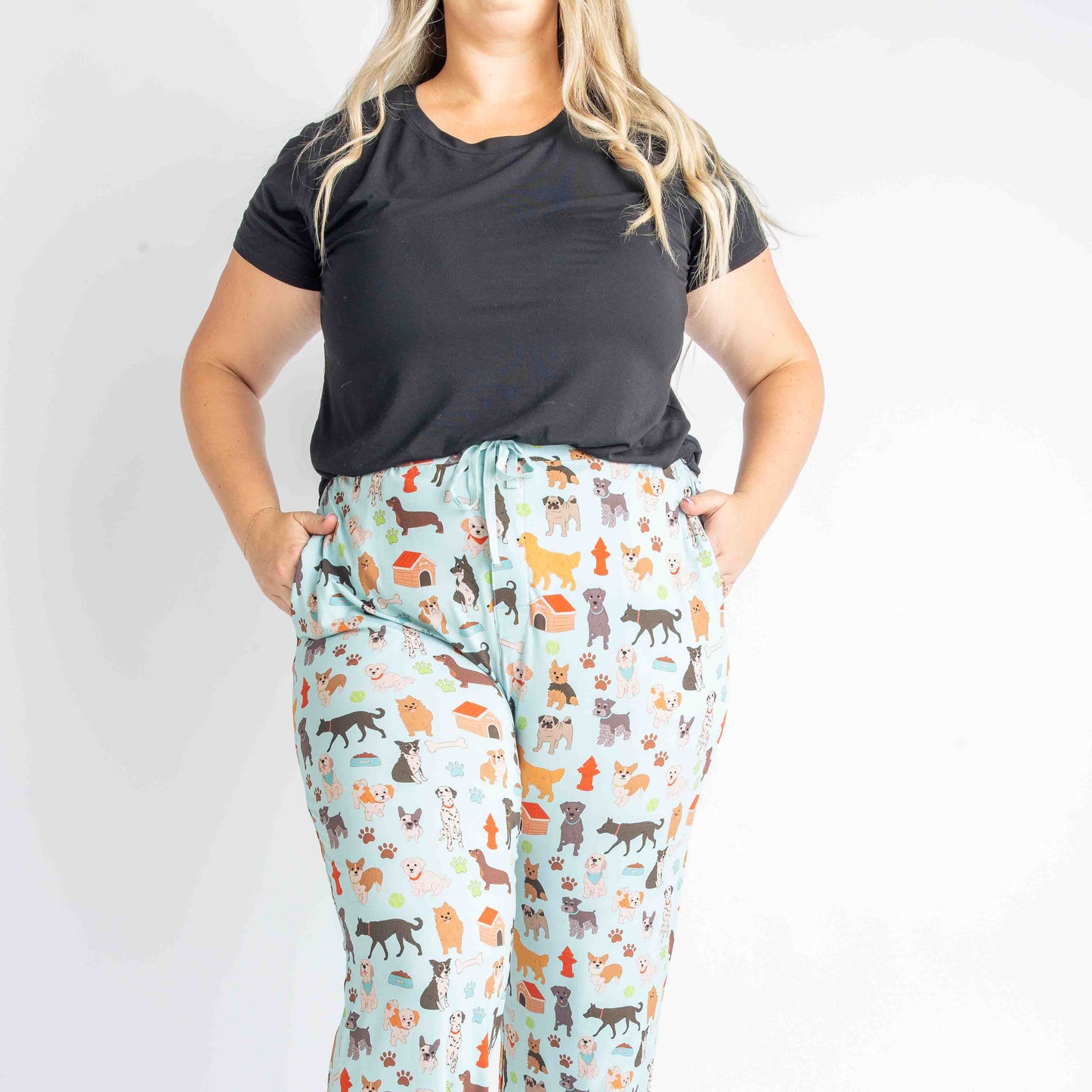 woman wears the "dogs bark" relaxed pants. she rest her hands in the pockets. she wears the solid black tee. the "dogs bark" print is a mix of various types of dogs, dog houses, dog bones, paw prints, dog food, and tennis balls scattered around a light blue background.