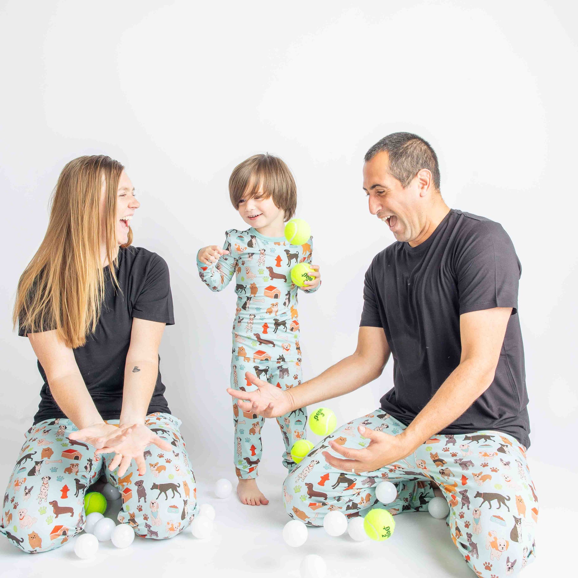 mom, dad, and child play with tennis balls. mom and dad are both wearing the "dogs bark" relaxed pants and solid black tee's. the child wears the "dogs bark" 2-piece matching set. the "dogs bark" print is a mix of various types of dogs, dog houses, dog bones, paw prints, dog food, and tennis balls scattered around a light blue background.