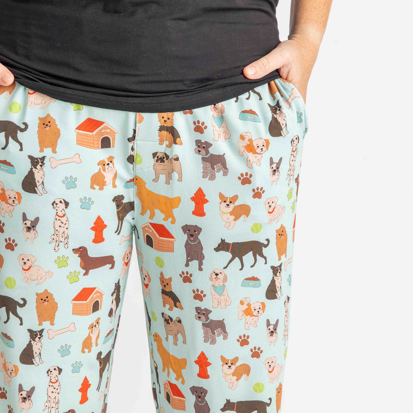 close up of a man wearing the "dogs bark" relaxed pants. he has his hands in his pockets. the "dogs bark" print is a mix of various types of dogs, dog houses, dog bones, paw prints, dog food, and tennis balls scattered around a light blue background.