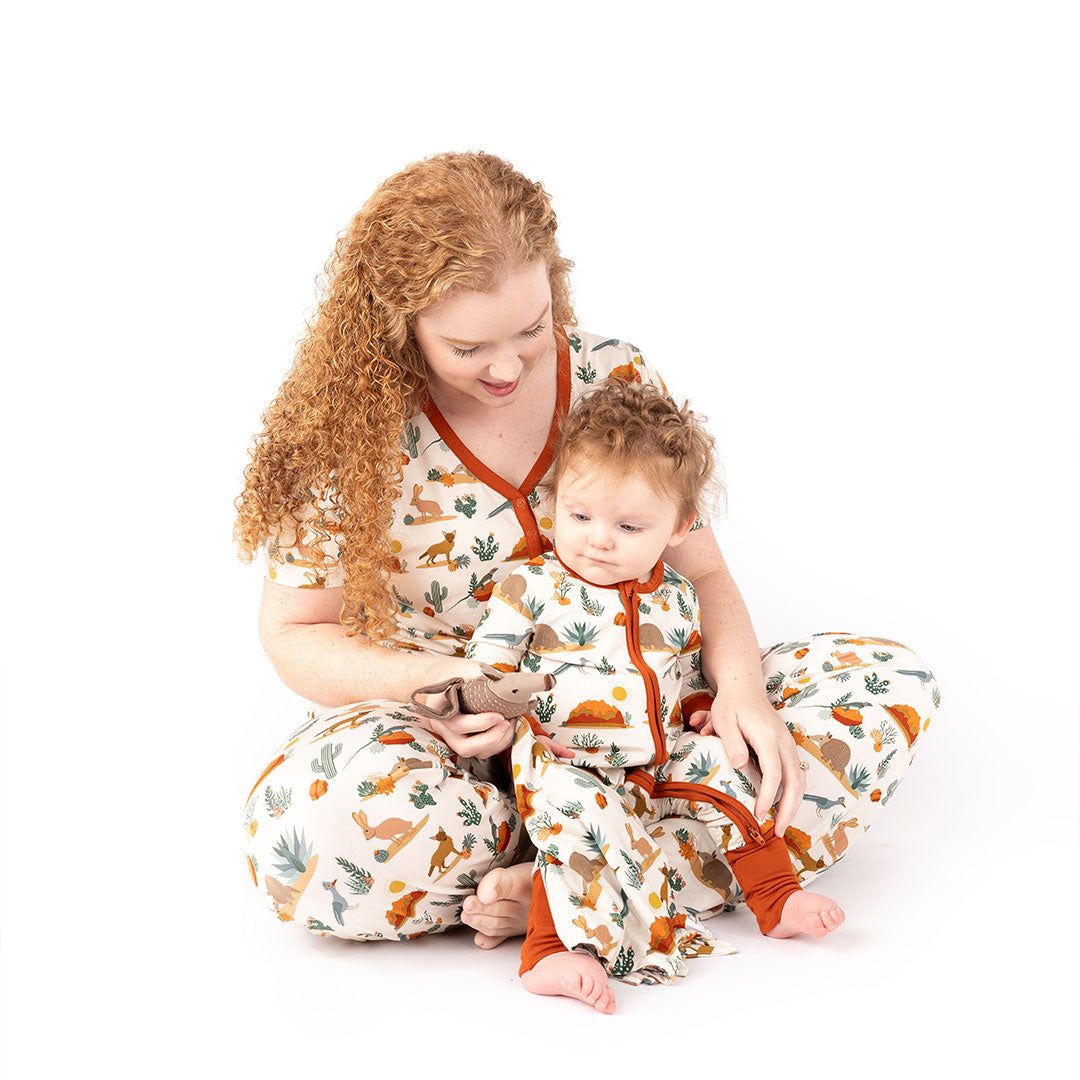 mom sits criss-sross in the floor with her baby in her lap. they are all wearing the "desert friends" print. the mom in the women's top and the relaxed pants. the baby in the convertible. they are playing with the lovey. the "desert friends" print is a scattered pattern of armadillo, cacti, other desert plants, rocks, desert foxes, lizards, and desert birds. this is all on a beige background. 