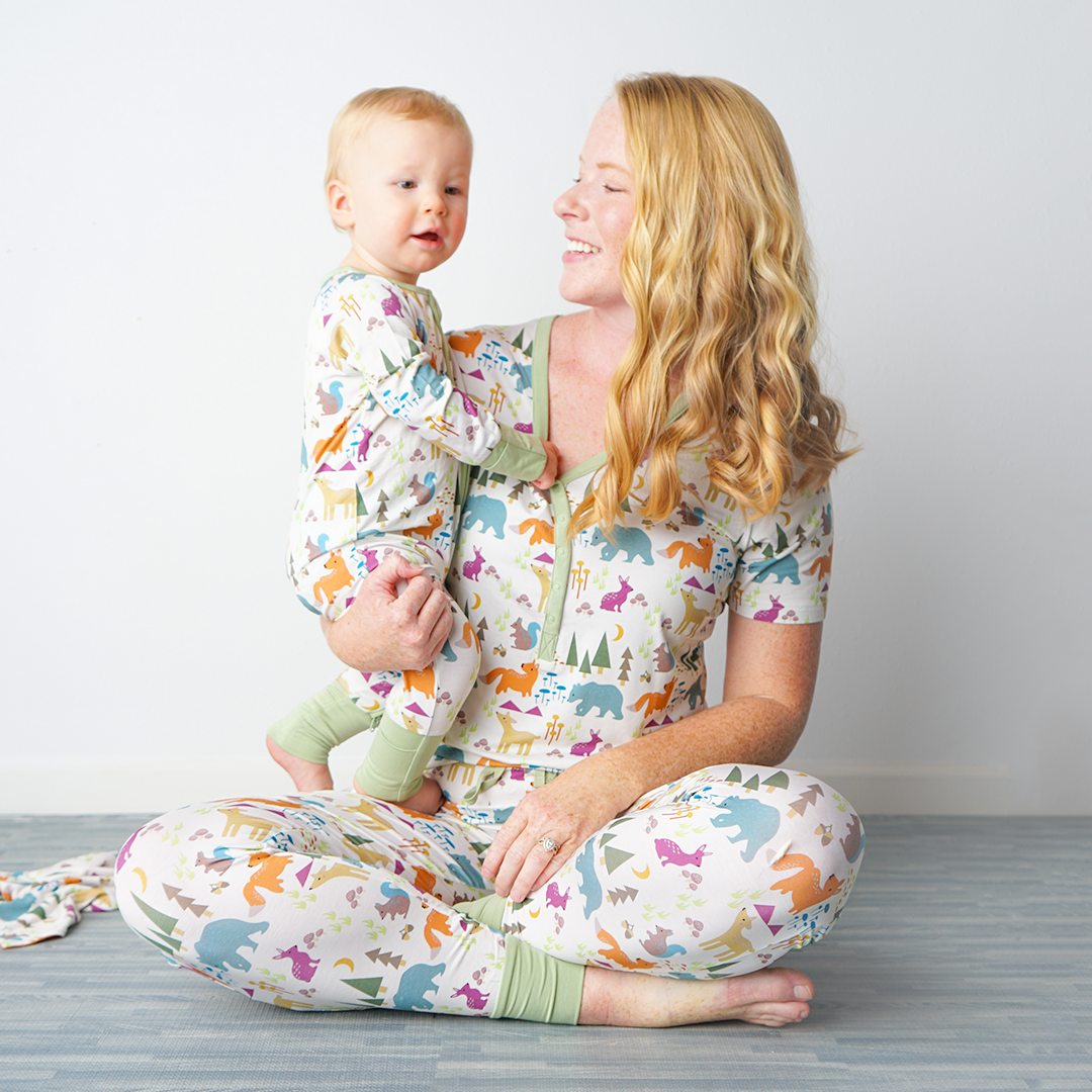 mom sits criss-cross on the floor and holds her baby up. the mom wears the "forest friends" women's top and joggers. the baby wears the convertible. the "forest friends" print has a mix of red foxes, brown deer, blue bears, purple bunnies, trees, grass, mushrooms, acorns, moons, and other colored dots scattered over a white background. 