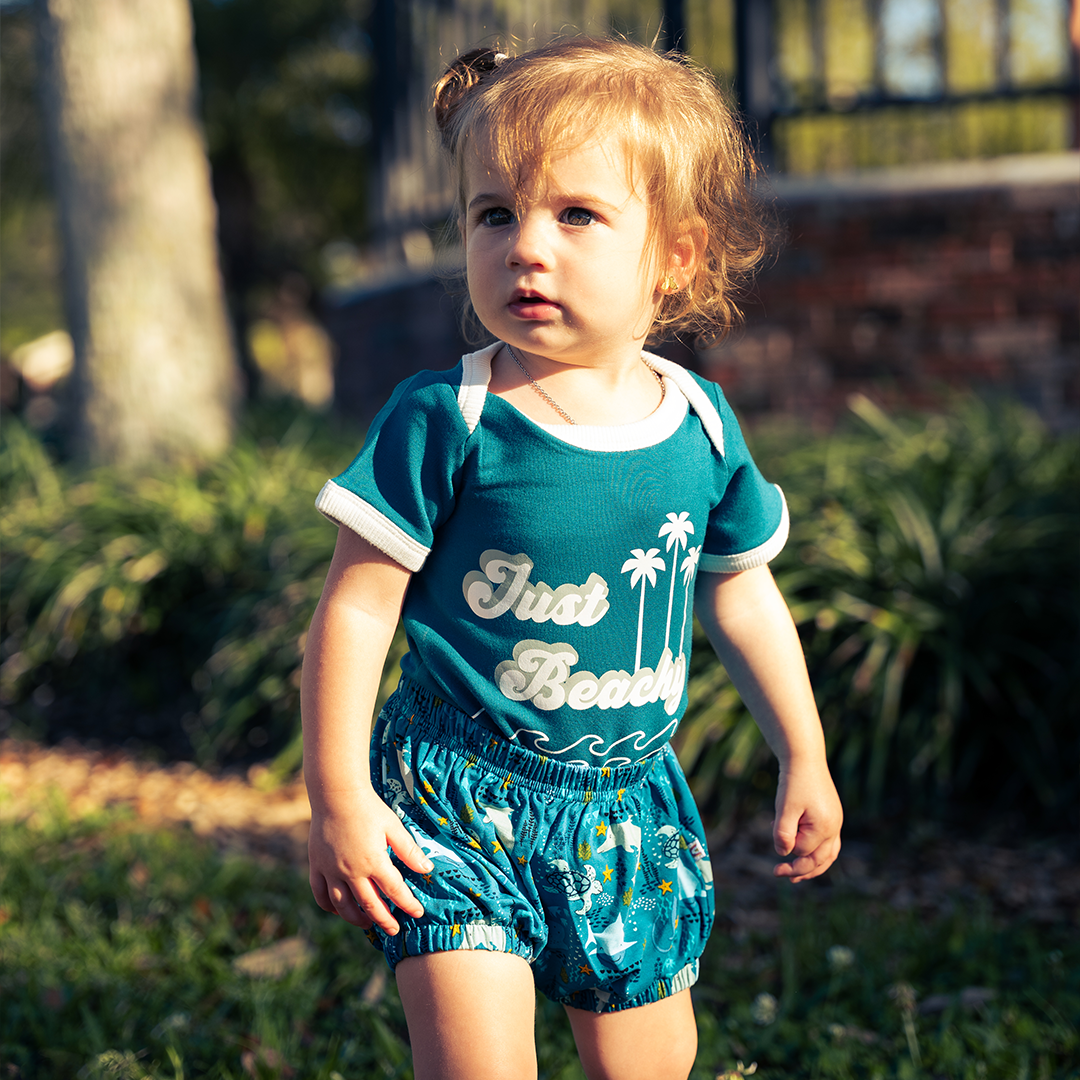 a little baby stands outside in his "ocean friends" baby bloomers and the "just beachy" onesie. the background is a blurred mix of a brick wall, bushes, and the trunk of a tree. the baby stands in grass. the "ocean friends" print is a combination of dolphins, stingrays, fish, starfish, coral, bubbles, and sharks, all spread out on a deep sea blue background. 