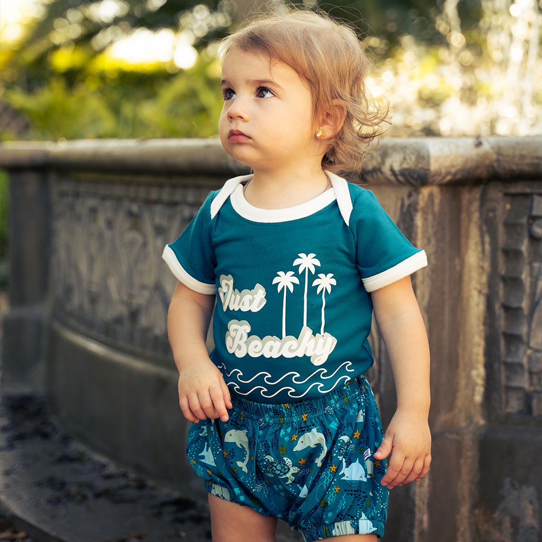 a little baby stands outside in his "ocean friends" baby bloomers and the "just beachy" onesie. the background is a blurred mix of a brick wall, bushes, and the trunk of a tree. the baby stands in grass. the "ocean friends" print is a combination of dolphins, stingrays, fish, starfish, coral, bubbles, and sharks, all spread out on a deep sea blue background. 