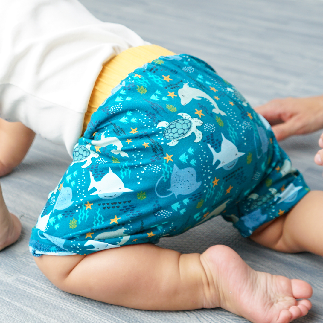 a focused picture of a baby crawling on the floor. he is wearing the "ocean friends" bamboo boy shorts. the "ocean friends" print is a combination of dolphins, stingrays, fish, starfish, coral, bubbles, and sharks, all spread out on a deep sea blue background. 