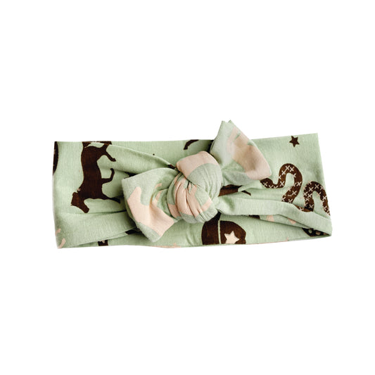 the "giddy up" baby headband with a bow on it. the "giddy up" print is a western look at horses, cowboy hats, boots, horse rings, cacti, moons and stars. colors on this print include green, brown, and white. 