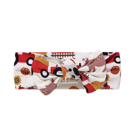 the "farm friends" baby headband, with a bow. the "farm friends" print is a collection of chickens, roosters, pigs, bails of hay, tractors, chicks, apples, and pumpkins. it's a white background and flashed of reds, yellows, and oranges, to bring out the farm aesthetic.