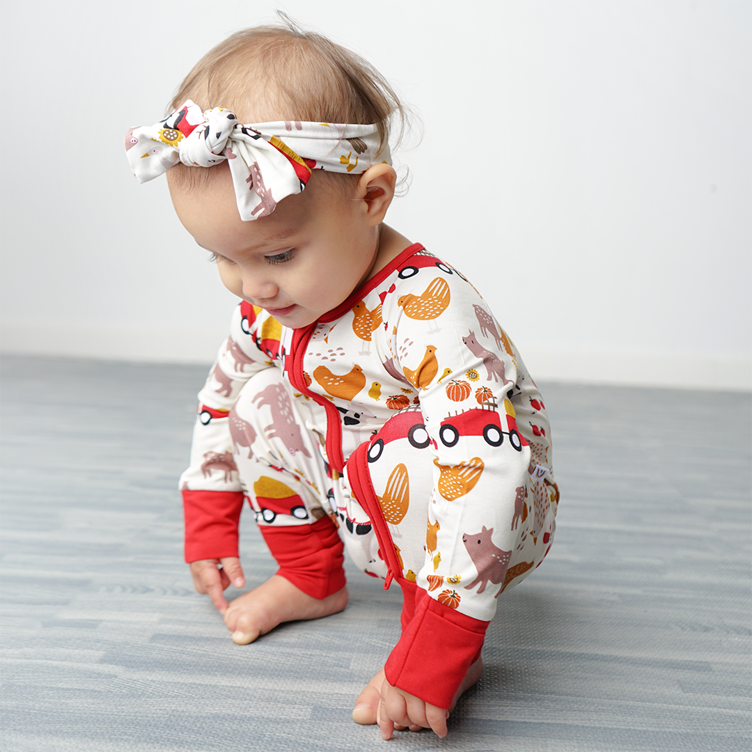 a baby girl squats on the ground and looks at the floor. she is wearing the print "farm friends" in the convertible and the headband. the "farm friends" print is a collection of chickens, roosters, pigs, bails of hay, tractors, chicks, apples, and pumpkins. it's a white background and flashed of reds, yellows, and oranges, to bring out the farm aesthetic.