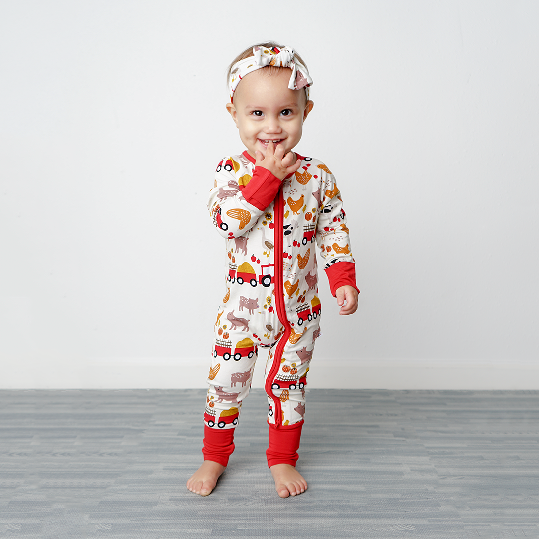 a small toddler girl smiles at the camera. she is wearing the "farm friends" convertible and the "farm friends" baby headband. the "farm friends" print is a collection of chickens, roosters, pigs, bails of hay, tractors, chicks, apples, and pumpkins. it's a white background and flashed of reds, yellows, and oranges, to bring out the farm aesthetic.