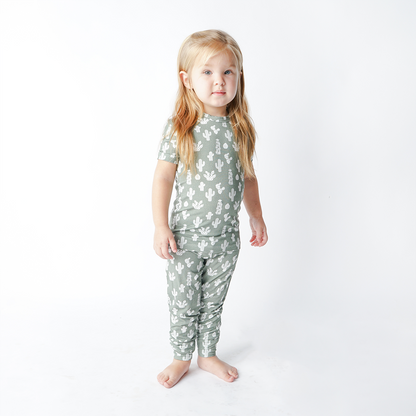 a young girl stands with her arms at her side. she wears the "stay sharp" matching pajama set. the "stay sharp" print is a variety of different white cacti on a greyish/green background. 