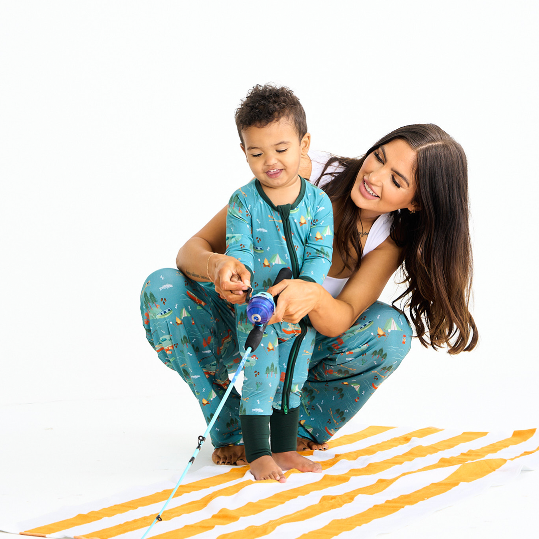 mom squats and stands behind her son. they play with a fishing pole. the boy is wearing the "jump in the lake" convertible, the mom wears the "jump in the lake relaxed pants. the "jump in the lake" pattern is a print to capture the day in the life on a lake. you can fine fishing poles, tents, camp fires, adirondack chairs, marsh mellows, ores, canoes, forest trees, and different types of fish and fishing gear.