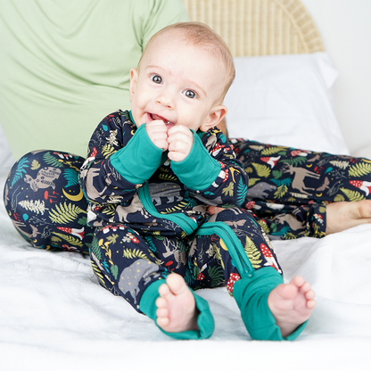 a small infant baby sits on his bottom. he his being help up by his parent's arm. the baby is smiling wide, with his hands to his mouth. the baby is wearing the "night forest" convertible. the baby and the parent are sitting on a bed. the "night forest" print is a night time themed design. you can see an array of forest animals ranging from, deer, bears, bunnies, birds, forest trees and leaves, flowers, and mushrooms. there are also starts and moons scattered around to enhance the night time atmosphere. 